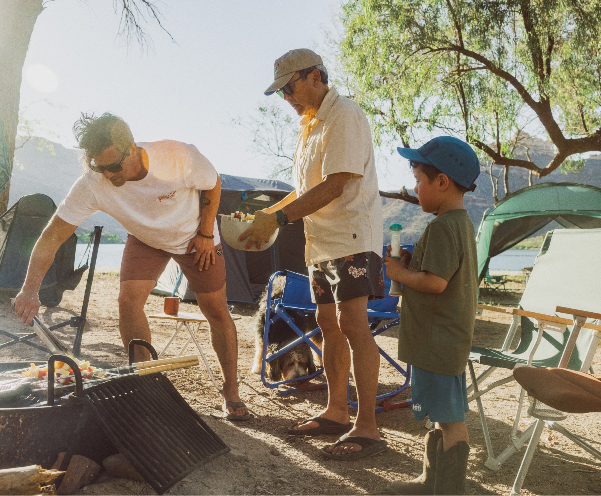 Three people gather around a fire pit to cook skewers.
