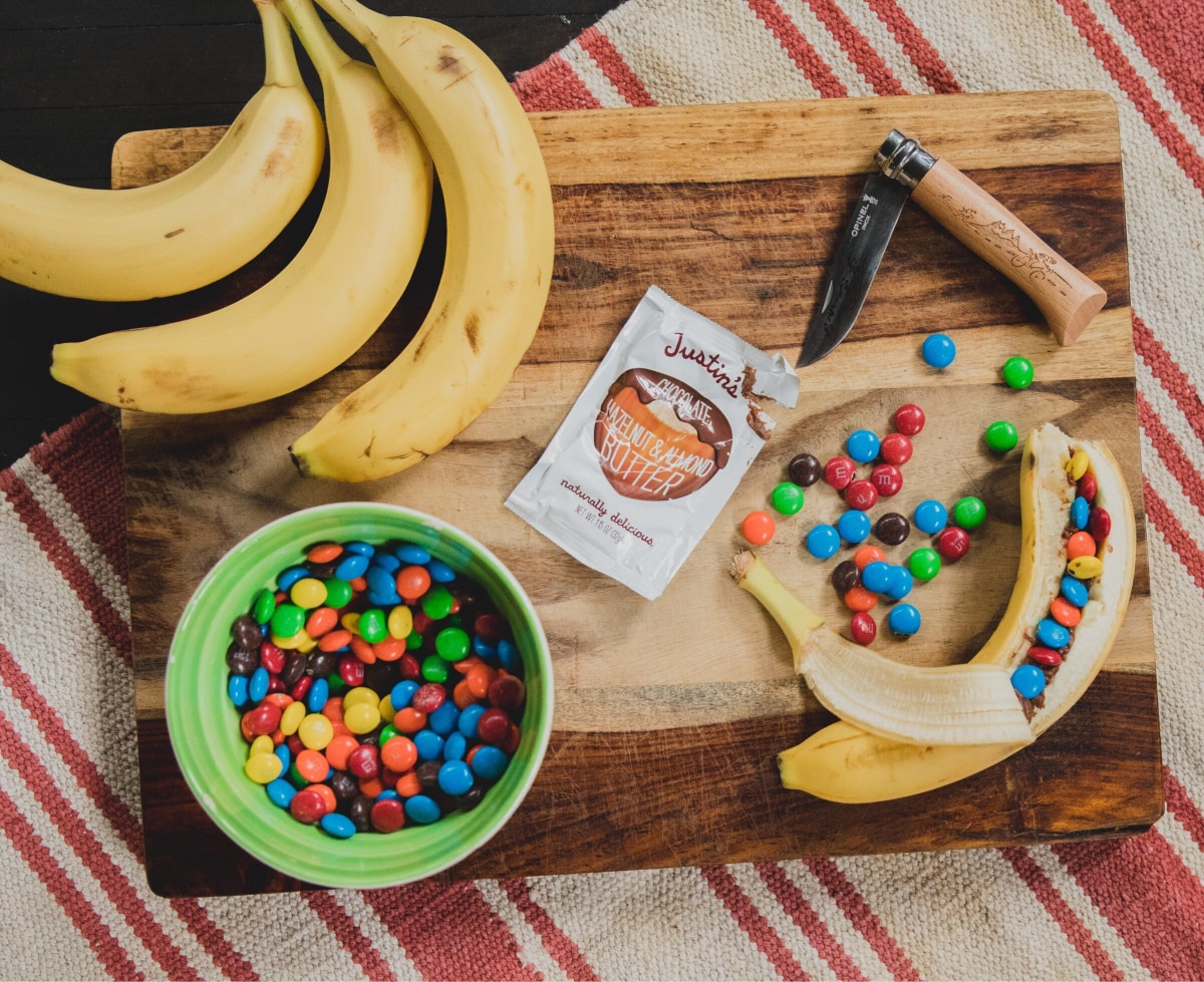Bananas, M&M’s, Justin’s hazelnut & almond butter, & an Opinel knife on a wood cutting board.