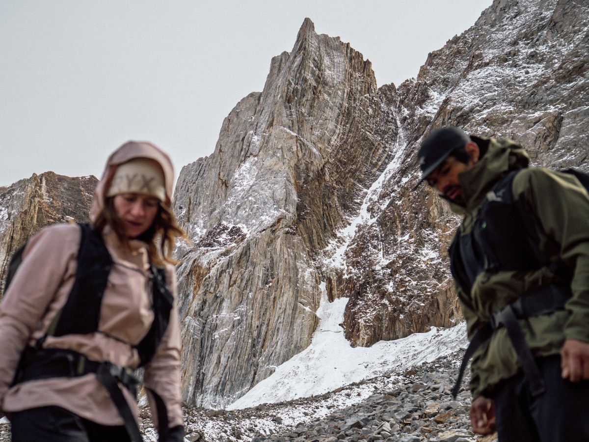 Two people look down while hiking with a snow covered rock formation in the background.