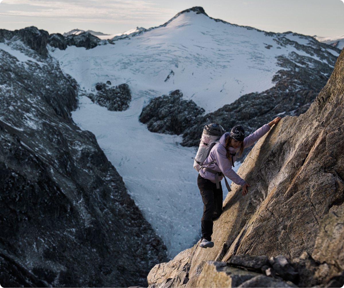 A person climbs a steep rock with snow covered mountains in the background.