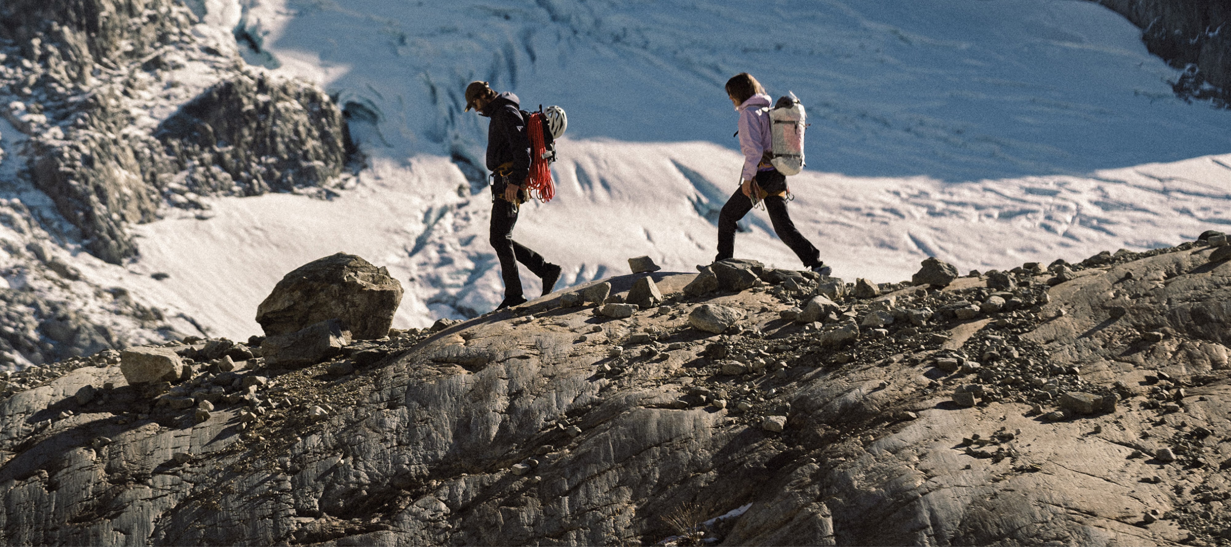 Two people hike over a large rock with snow covered mountains in the background.