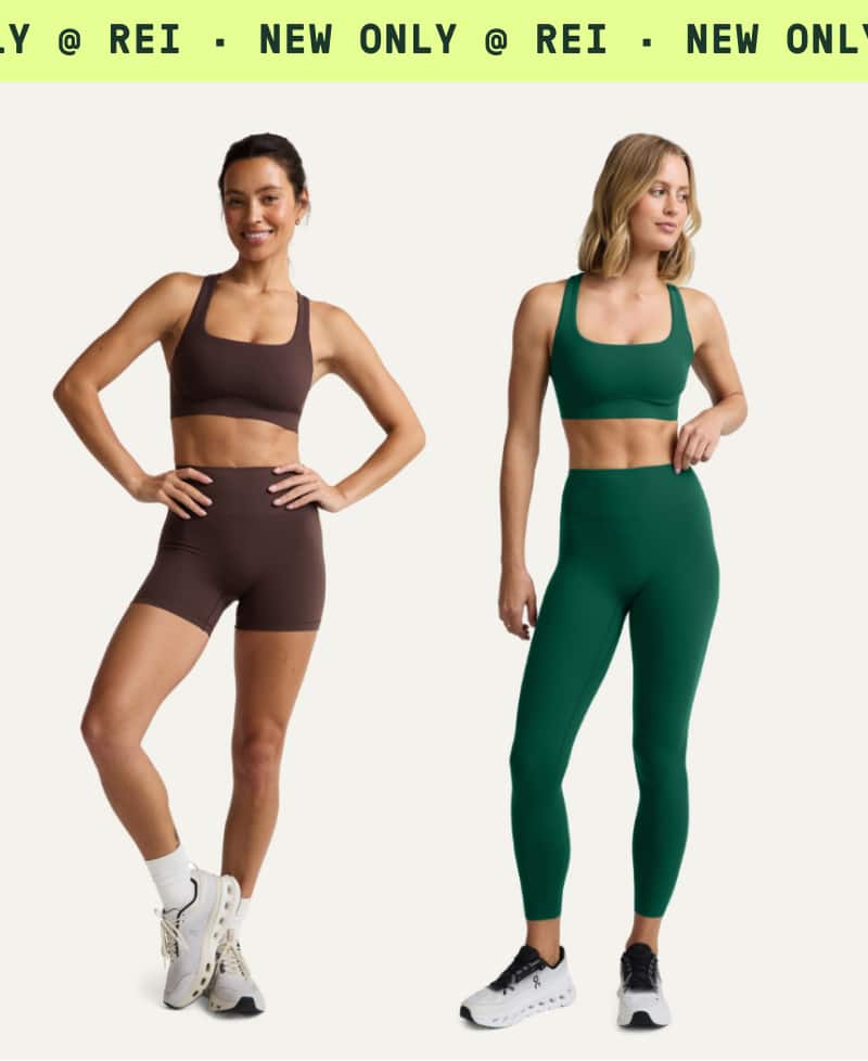 Two people pose in Beyond Yoga clothing on a light colored background. Ticker tape text reads: ‘New only at R E I.’