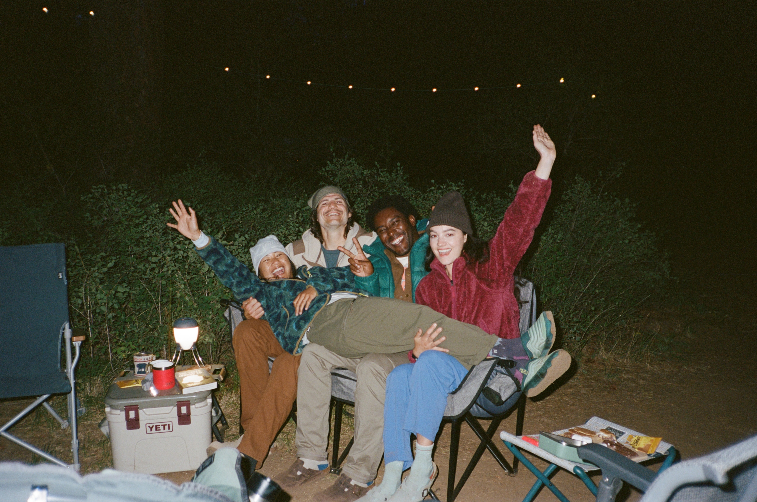 A group of three smiling people on camp chairs at a campsite have a fourth person stretched out across their laps.