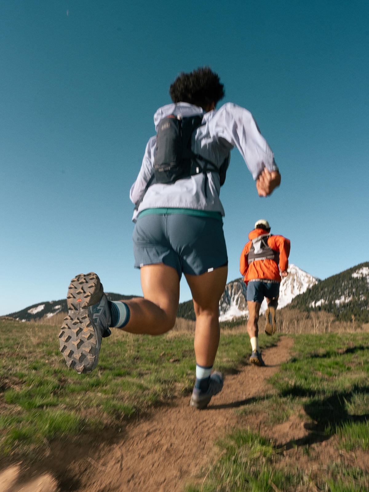 Two people run through a dirt trail towards large mountains.
