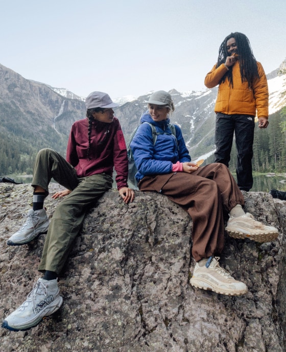Three hikers stop to sit down on large rocks with a view of mountains in the background.