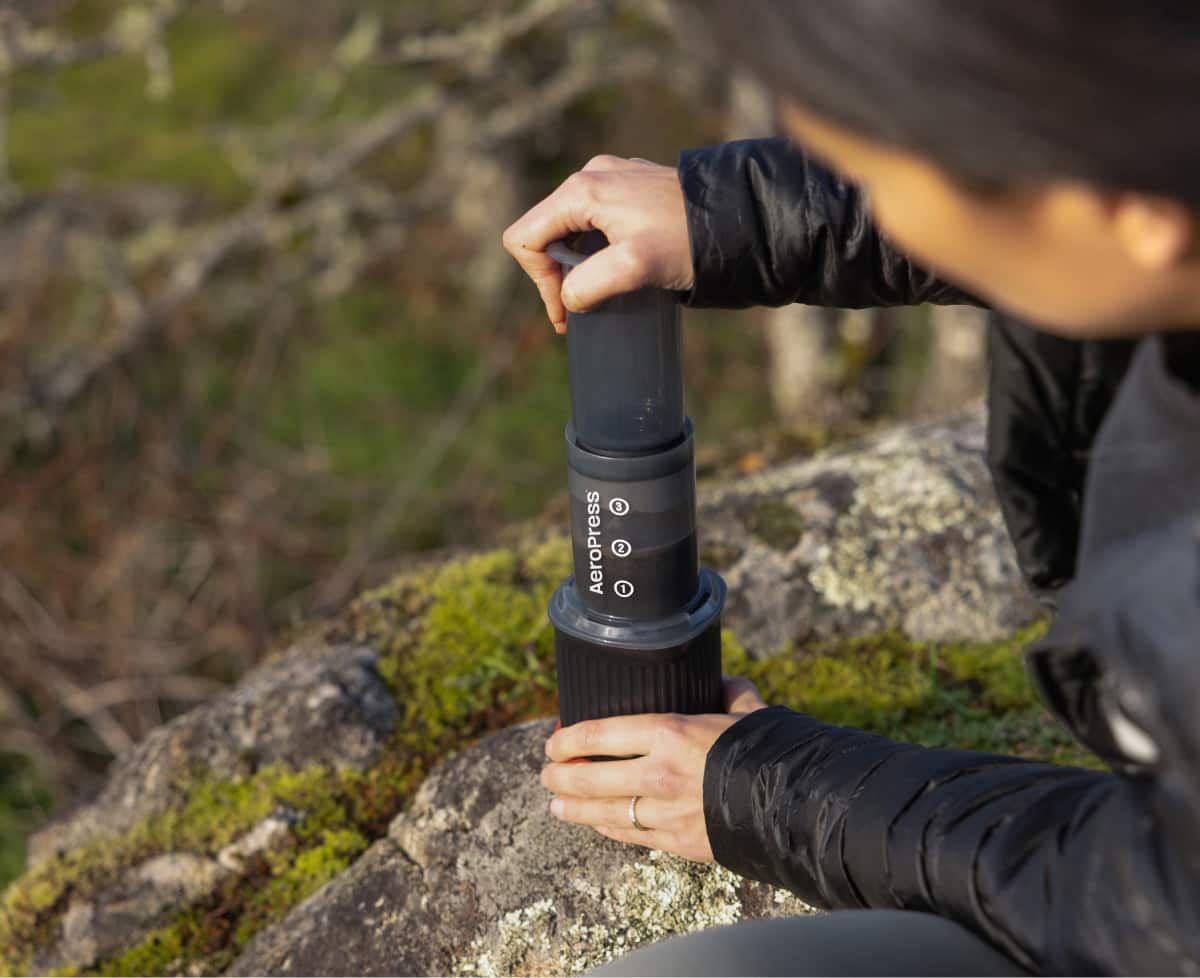 A person uses an AeroPress to make coffee at camp.