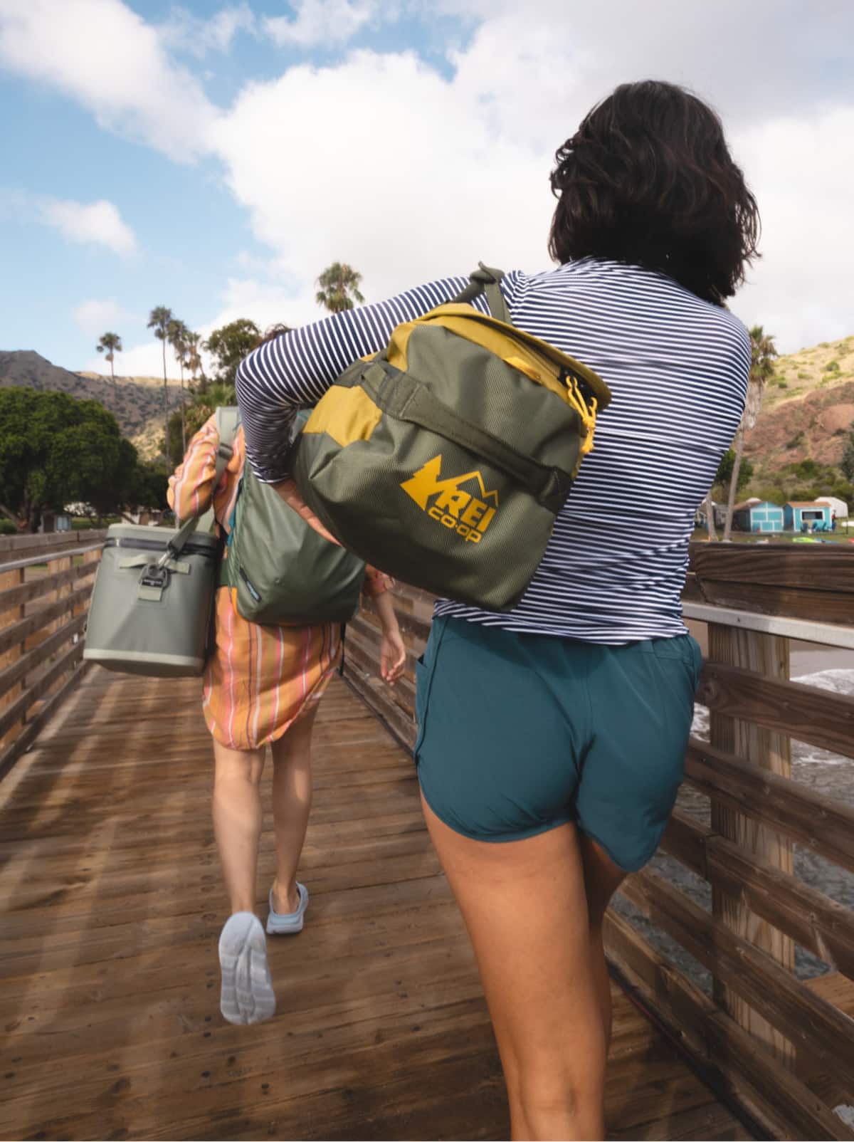 Two people walk across a wooden bridge with their bags.