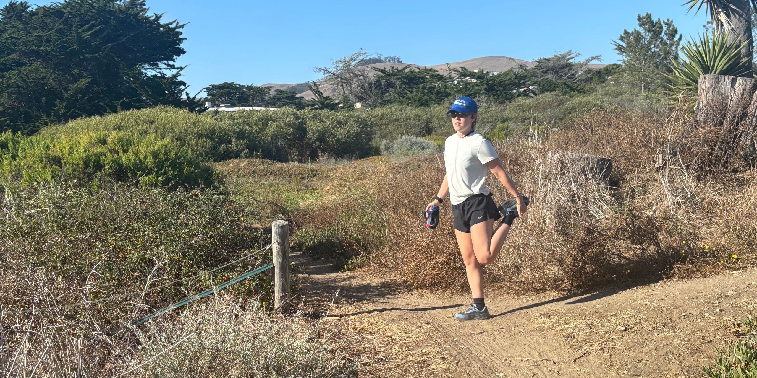 A person in a blue hat, white shirt and black shorts stretching their quad while holding a water bottle on a trail.