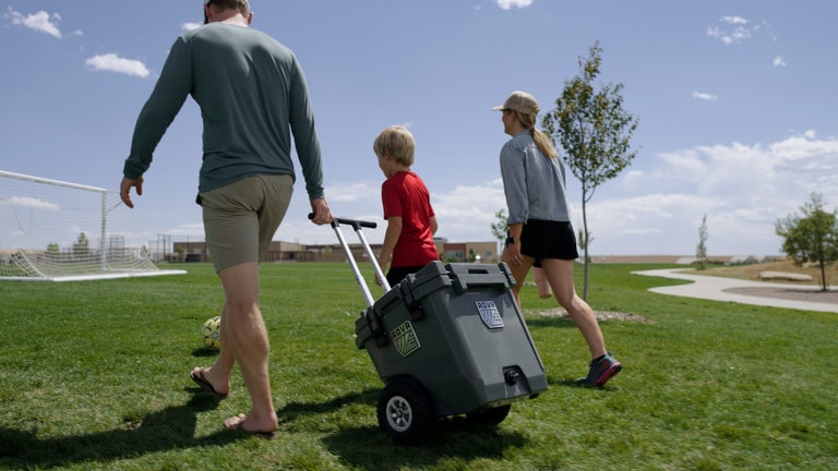 Three people walk across a lawn with one pulling a wheeled cooler.