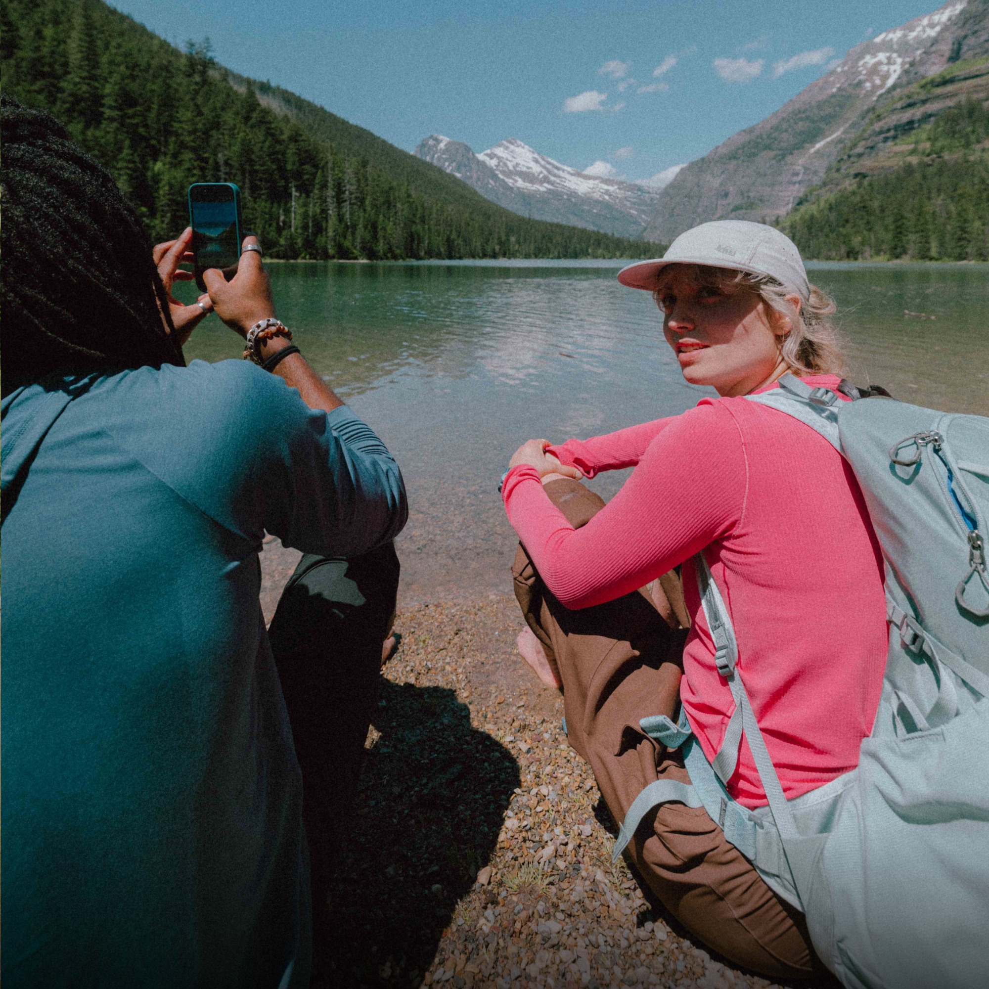 Two hikers sit down to relax near a lake with a view of large mountains in the background.