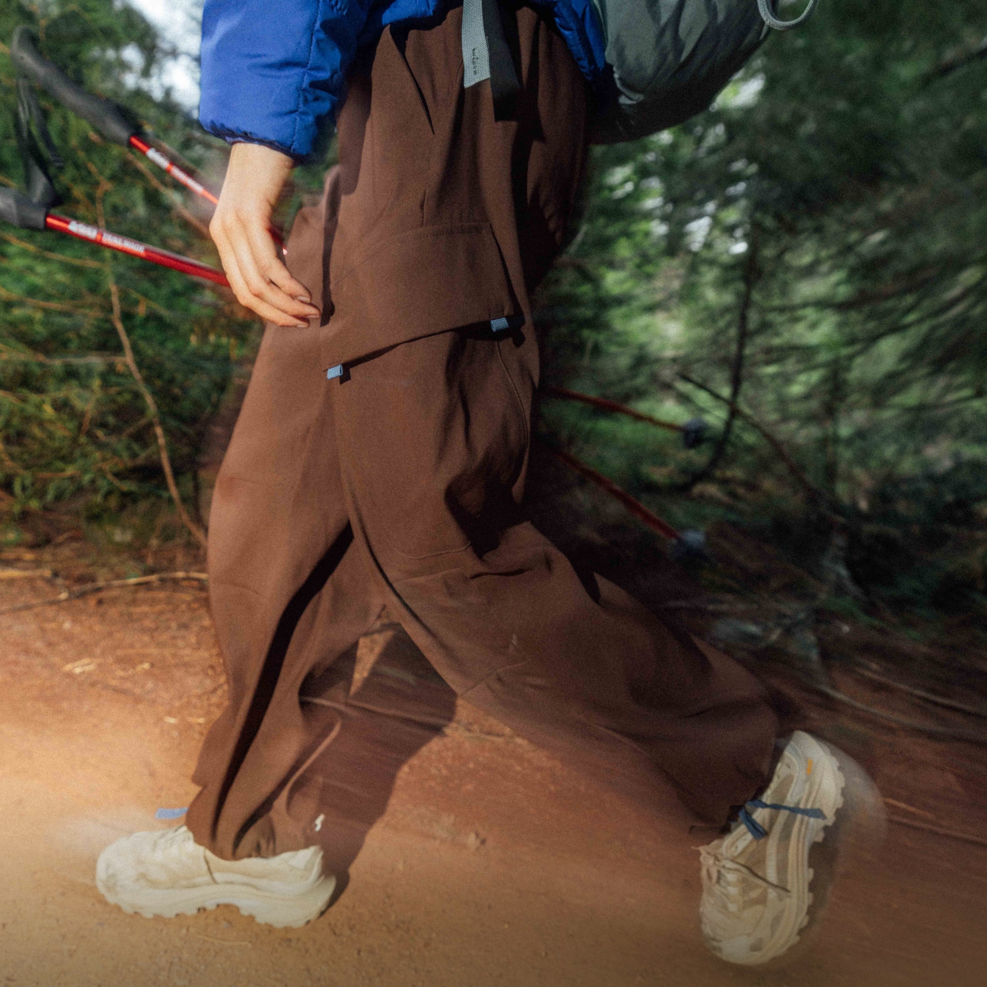 A person hikes through a dirt trail with a backpack on and trekking poles in their hand. 