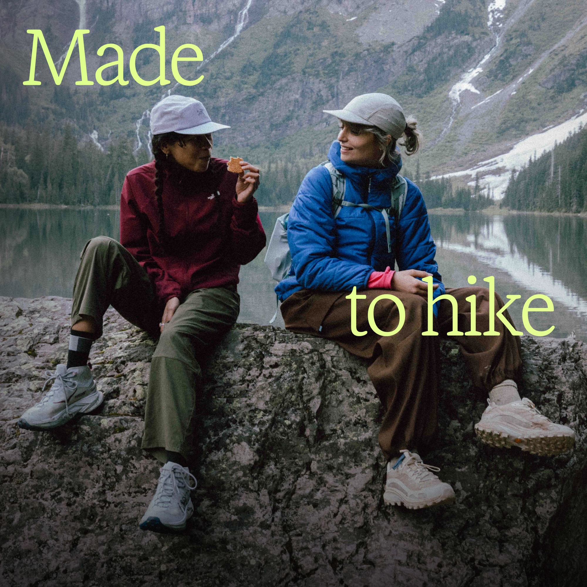 Two hikers sit on a rock together with a lake and large mountains in the background.