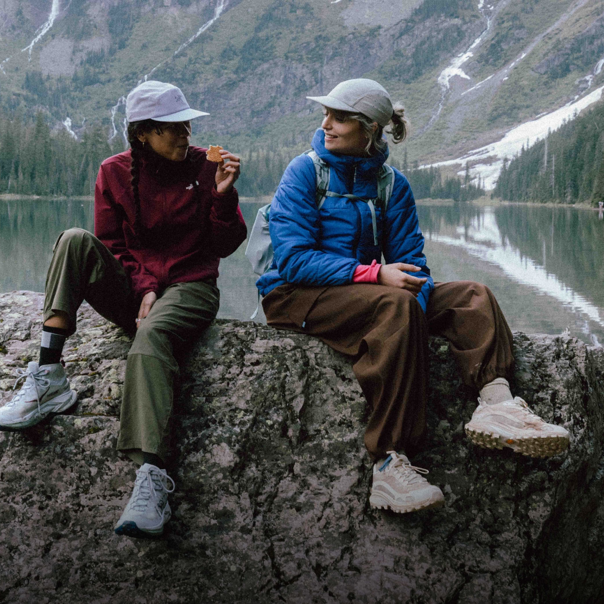 Two hikers sit on a rock together with a lake and large mountains in the background.