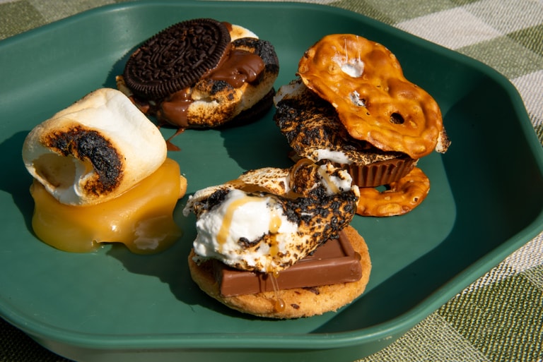 A green plate with four unique sweet s'mores on it, including a peanut butter cup and pretzel, an Oreo cookie, and more
