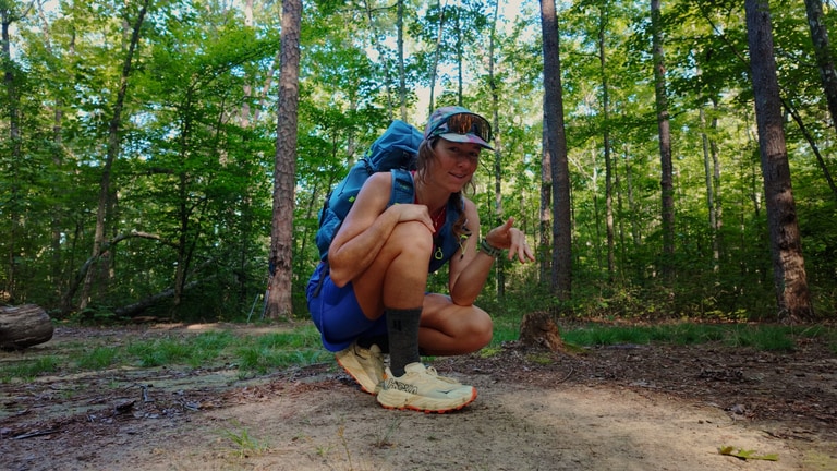 A woman kneeling on a trailing, wearing fastpacking gear including a blue backpack and white and yellow trail-running shoes.