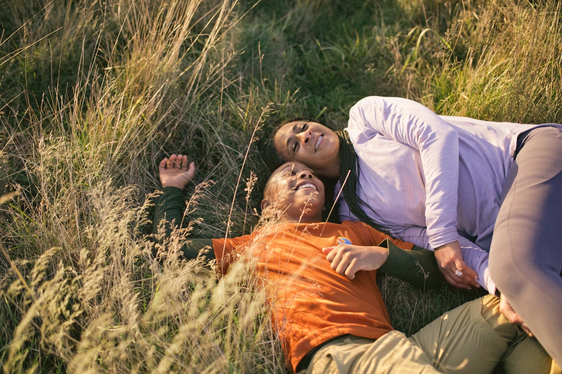 A smiling mom and her young son lay outdoors, relaxing on a field of grass in the warm afternoon sunshine.
