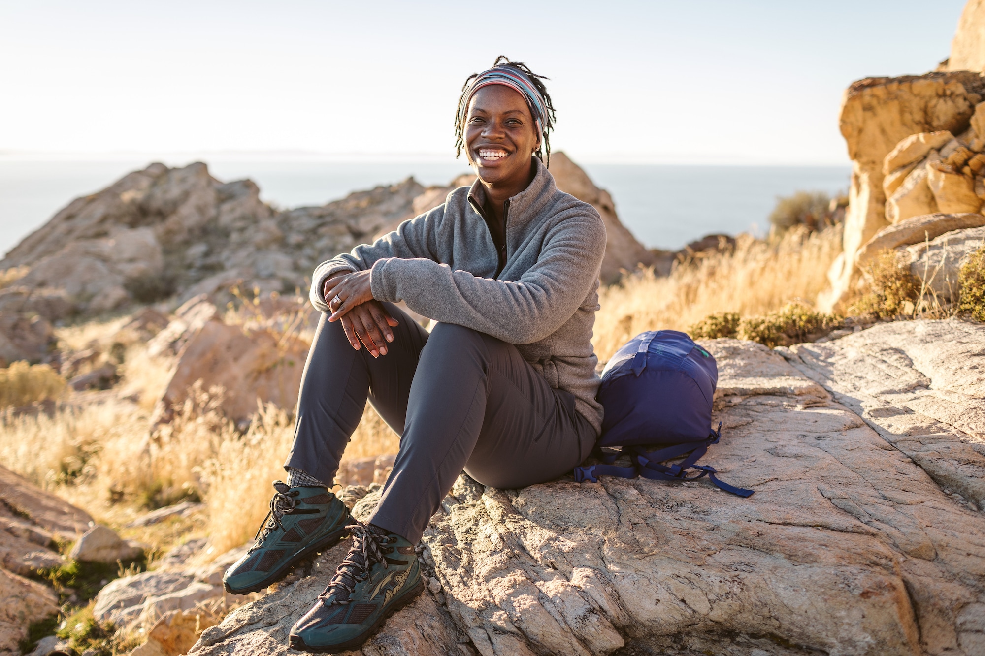 A smiling woman wearing midweight outdoor athletic attire sits on a rock amidst a landscape of rock, scrub brush, and clear blue sky