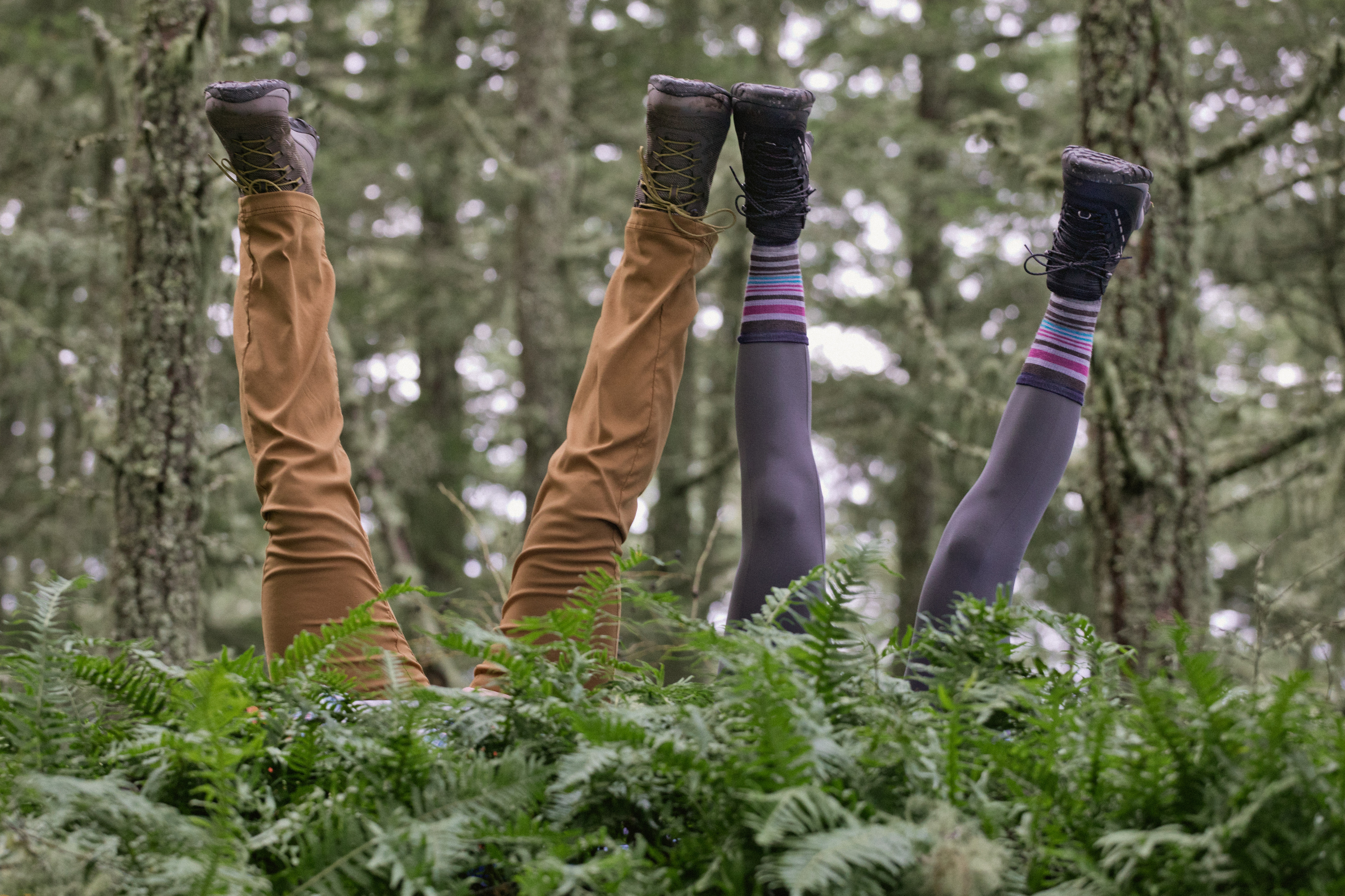 Against a forest backdrop, with bright green ferns in the foreground, two pairs of legs dressed in midweight outdoor attire and hiking boots stick up in the air, implying two people behind the ferns doing headstands