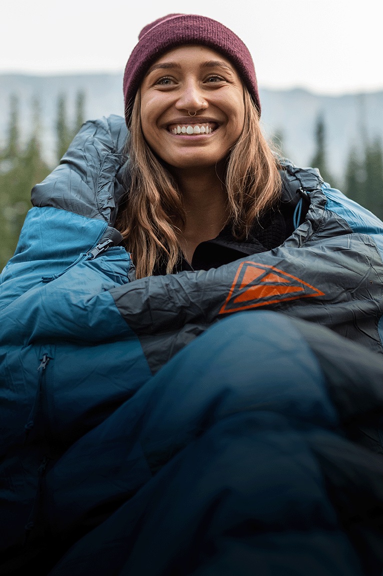 A camper in the Kelty Cosmic 20 sleeping bag sits on the ground and holds a mug