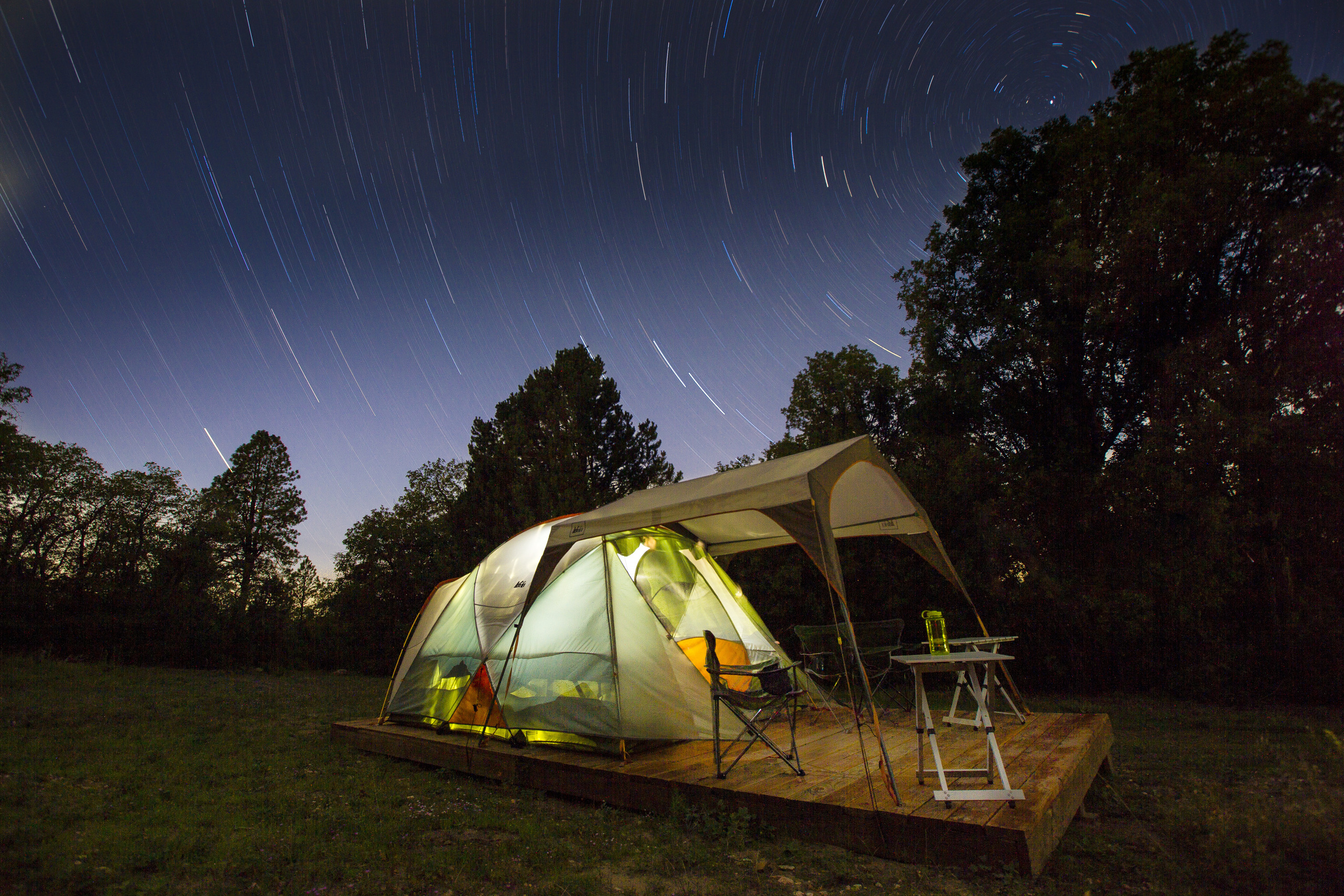 A tent with an extended shelter top is illuminated at night under a vast night sky of stars.