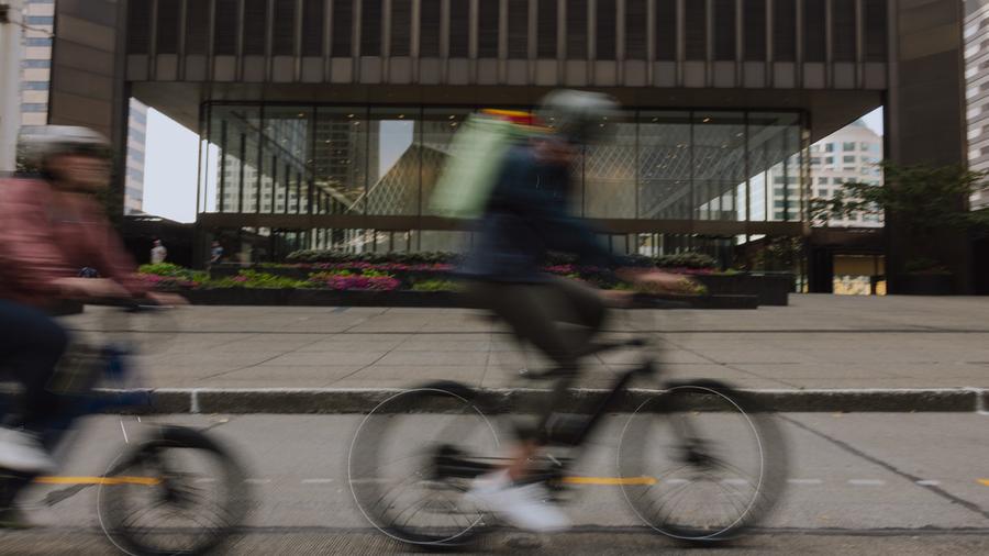Two people pedal e-bikes along a road in front of a building
