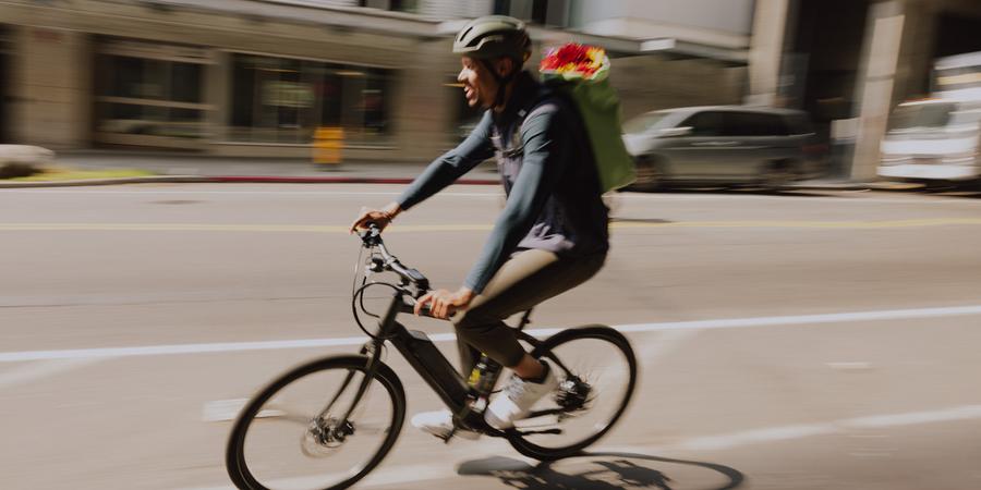 A person carrying flowers in a backpack pedals an electric bike through town