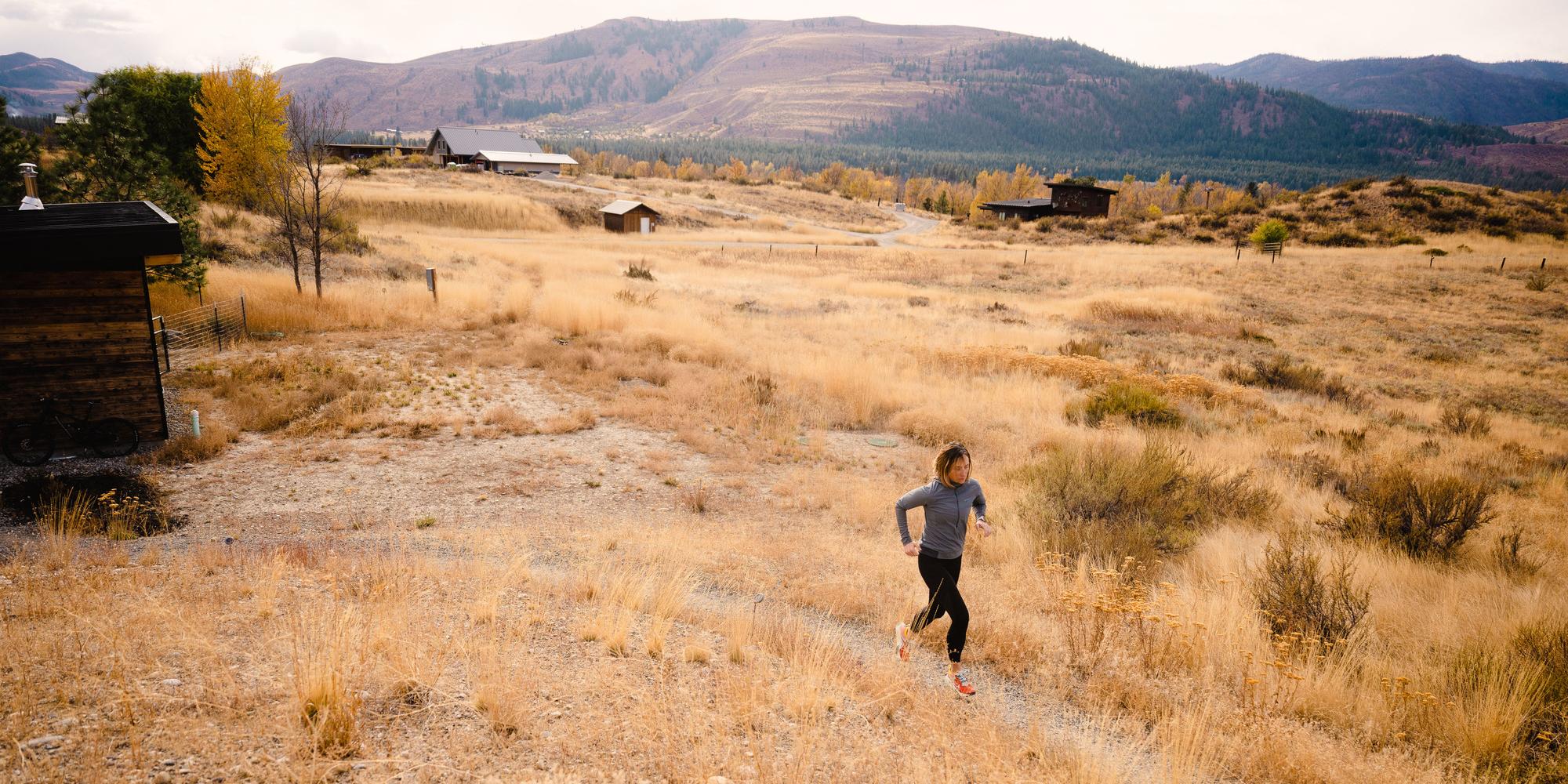A runner traverses a trail with a large mountain range behind her and a small cabin to her right.