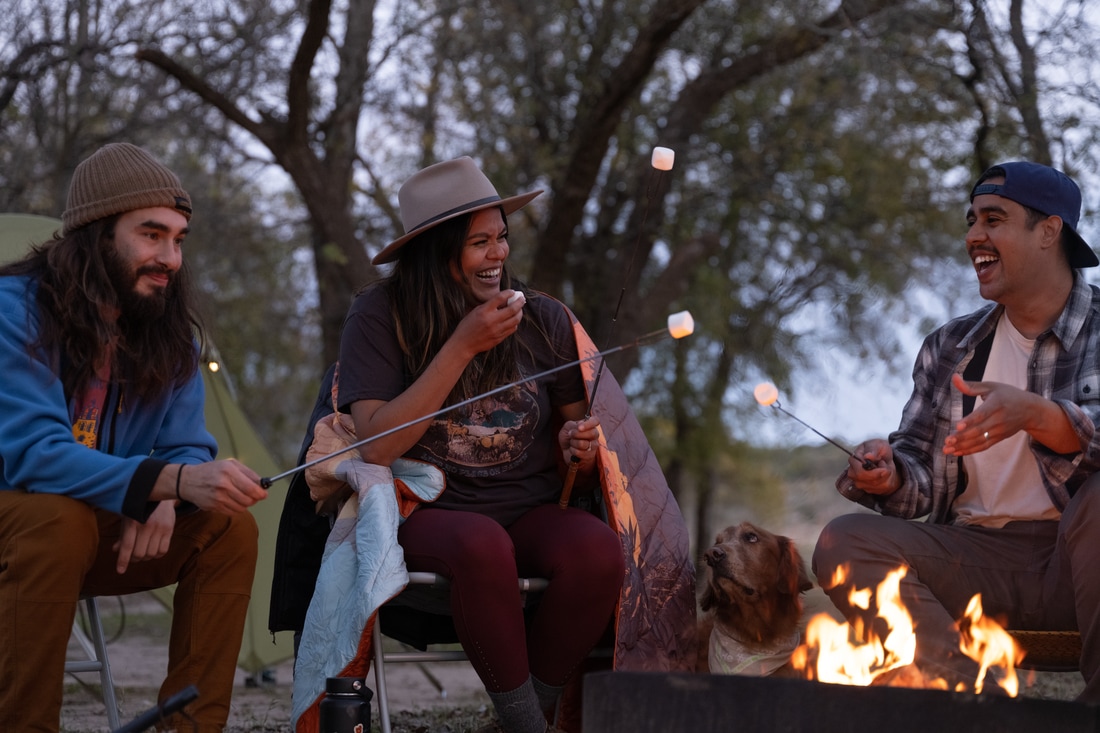 Three friends and a dog sit around a campfire, laughing and roasting marshmallows as the sun sets.
