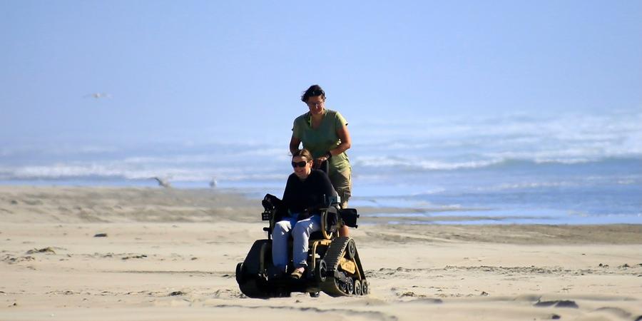 Person using a track chair on a beach with a companion walking behind her