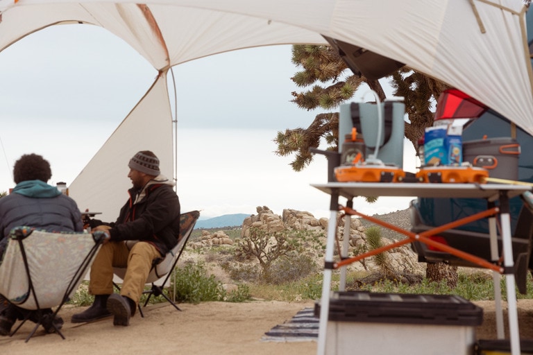 Two people sit under a camp shelter near a camp kitchen table