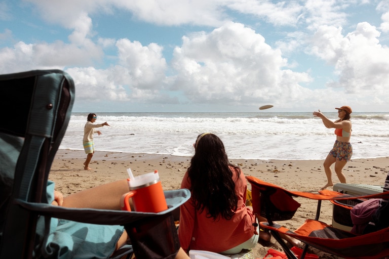 While people play on the beach, a person sits in a beach chair with a water bottle nestled in the chair pocket.