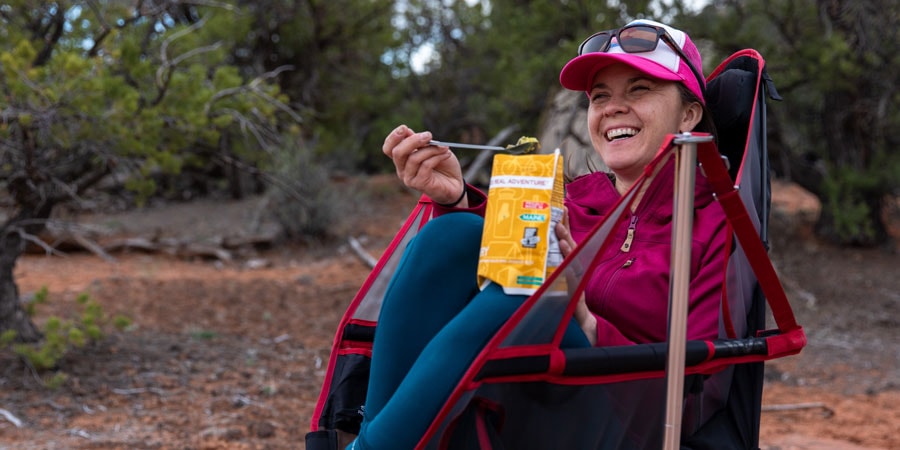 Smiling person sits in a camp chair holding a spoon and eats from a dehydrated meal in a beautiful outdoor setting.