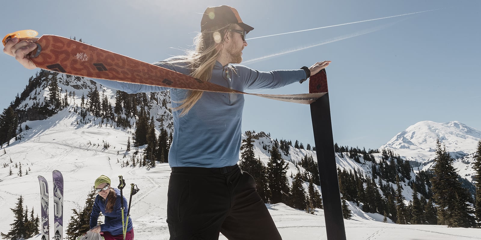 Skier standing at the base of a mountain pulling a climbing skin off the bottom of his ski. Fellow skier in the background.