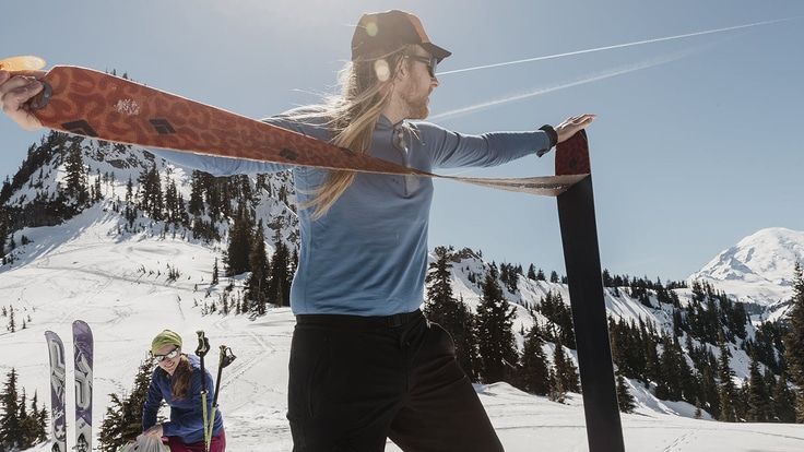 Skier standing at the base of a mountain pulling a climbing skin off the bottom of his ski. Fellow skier in the background.