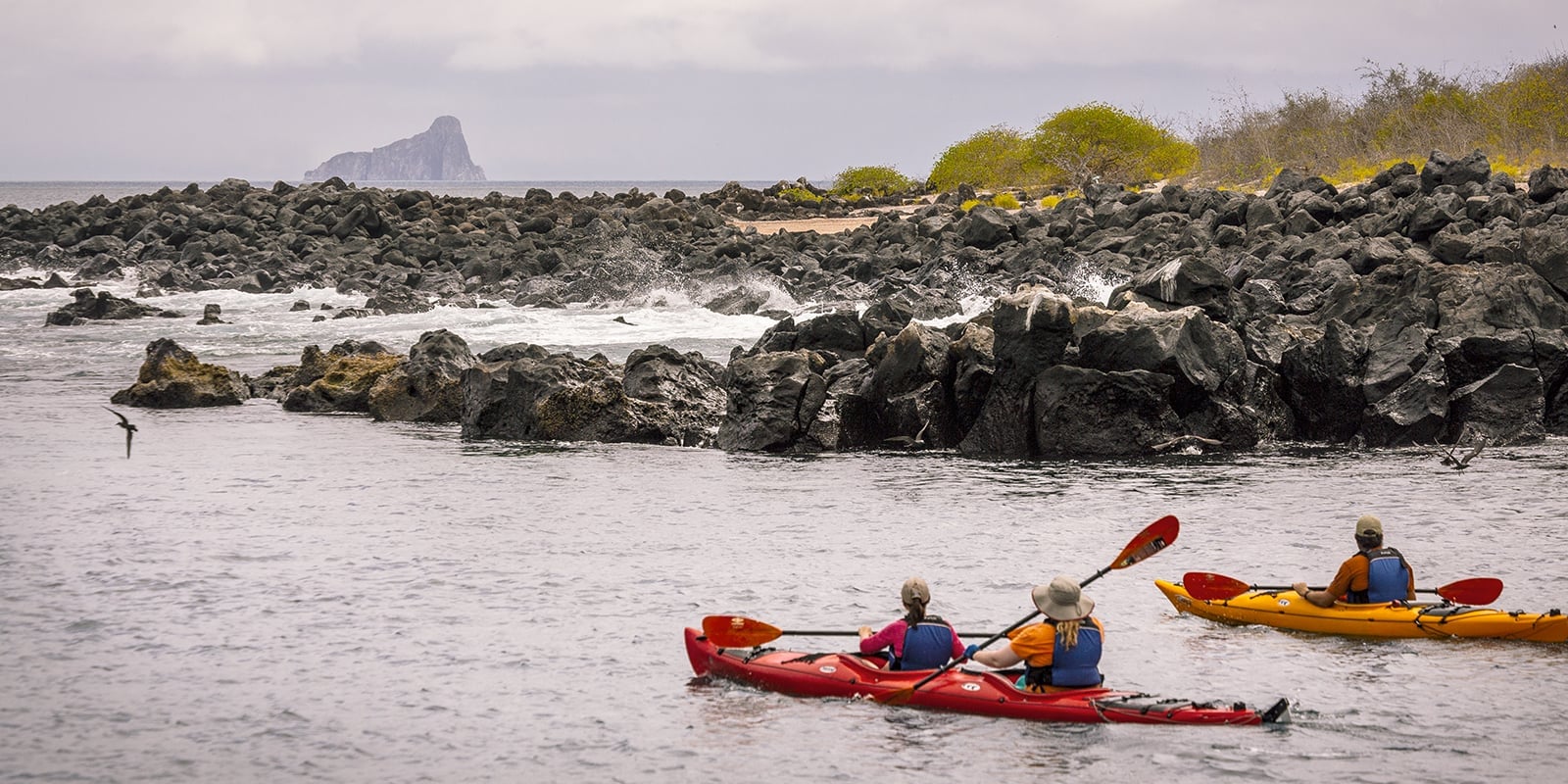 Two kayaks approach a rocky section of a beach.  In the distance there is an island made of steep rock cliffs.