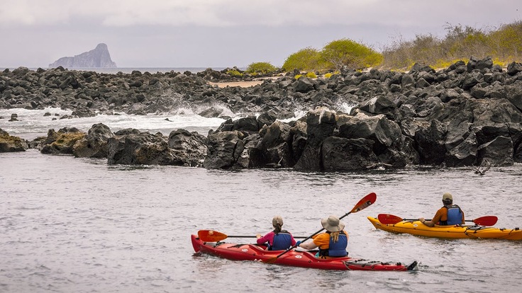 Two kayaks approach a rocky section of a beach.  In the distance there is an island made of steep rock cliffs.