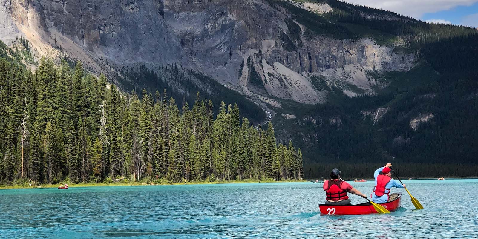 Two people canoeing on a mountain lake with fir trees on the far shore.