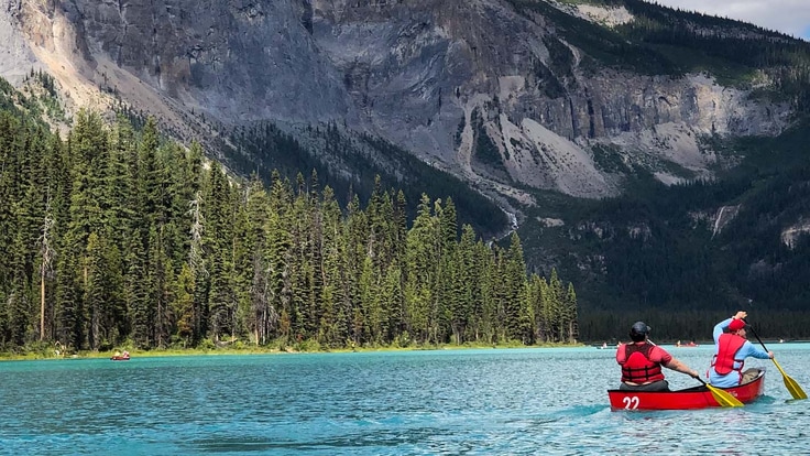 Two people canoeing on a mountain lake with fir trees on the far shore.