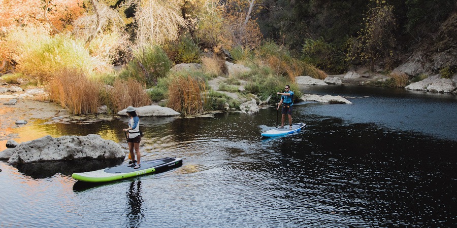 Two stand up paddle boarders make their way along an inky-blue flat-water river.
