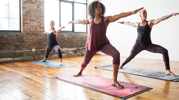 3 women doing the warrior 2 yoga pose on mats in a studio with hard wood floors and concrete walls.