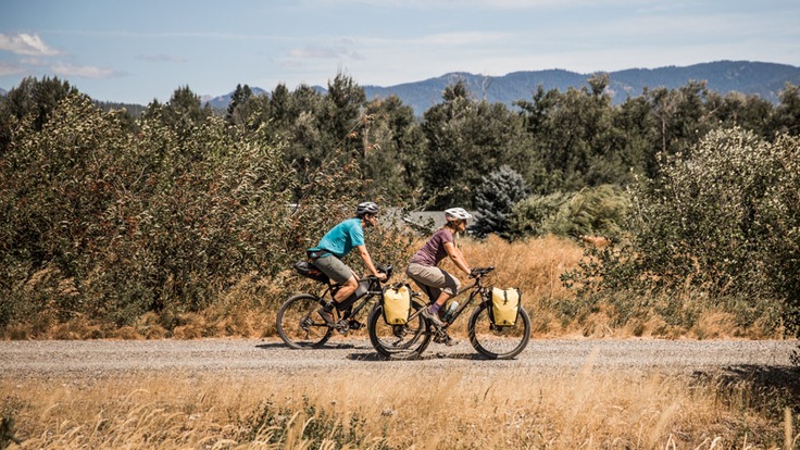 A man and woman on a bikepacking trip ride their bikes down a gravel road