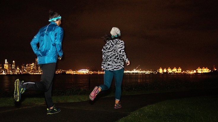 Two runners, each wearing reflective gear and a headlamp, striding on a path toward distant city lights.