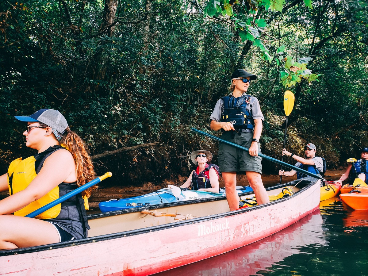 A guide stands on a canoe while other paddle in kayaks down a river
