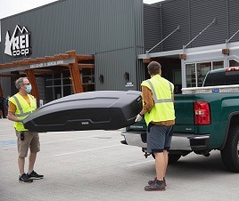 Two R E I employees load a roof box into the back of a customer's pickup truck.
