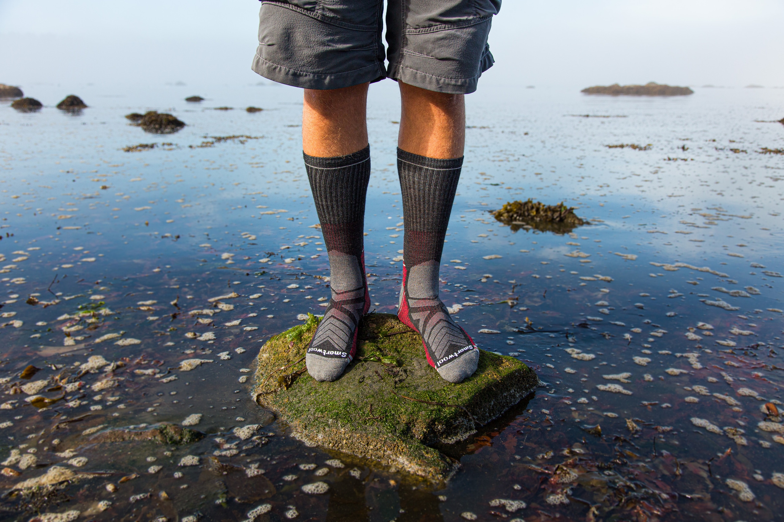 A person stands on a moss-covered rock in their sock feet, surrounded by marshy water.