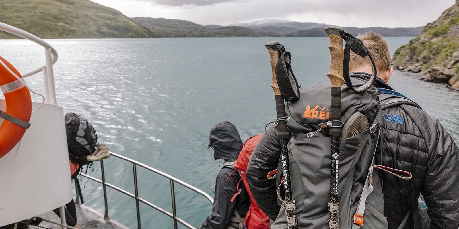 People dressed in hiking gear on the edge of a boat looking out over water, with land in the distance.