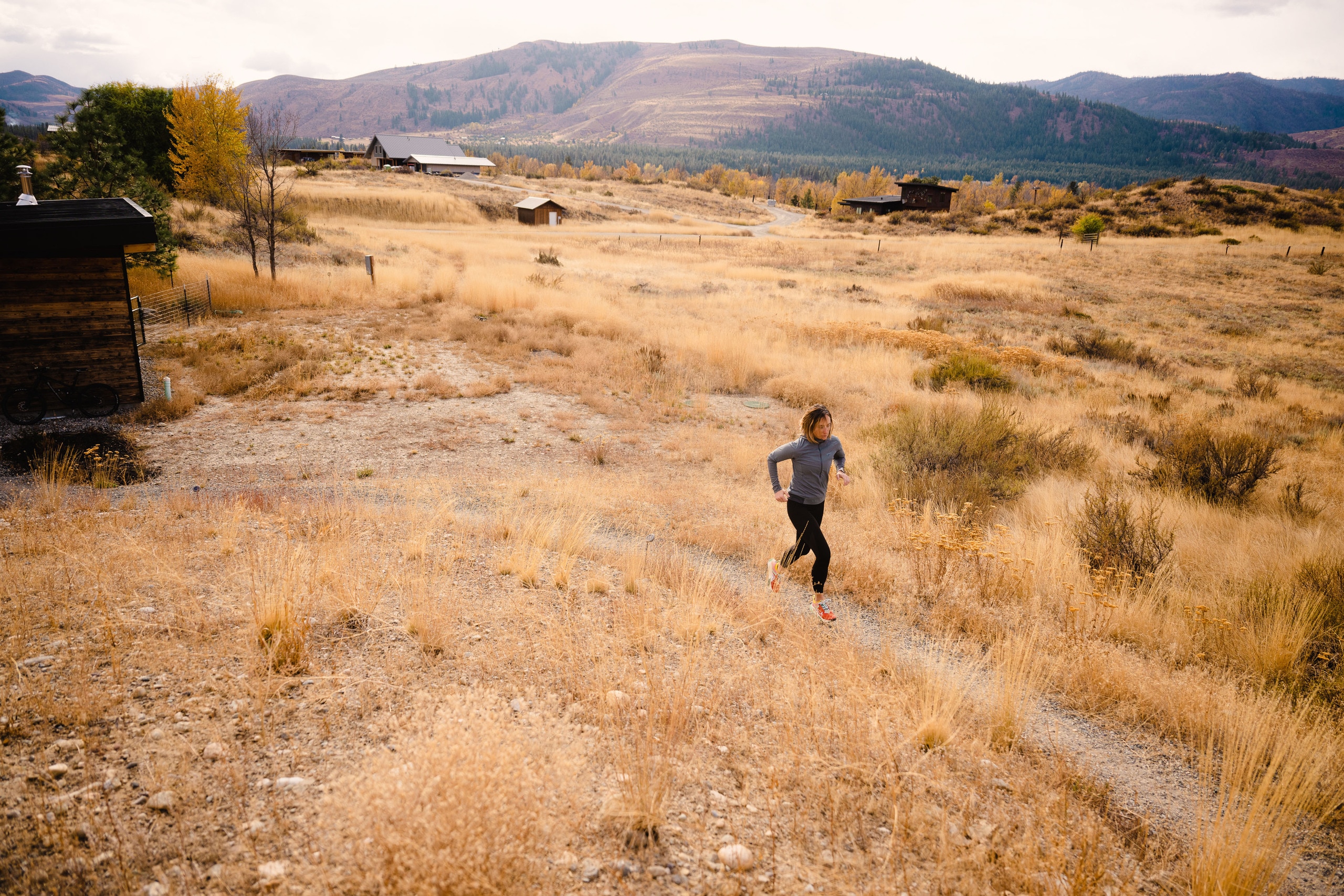 A runner traverses a trail with a large mountain range behind her and a small cabin to her right.