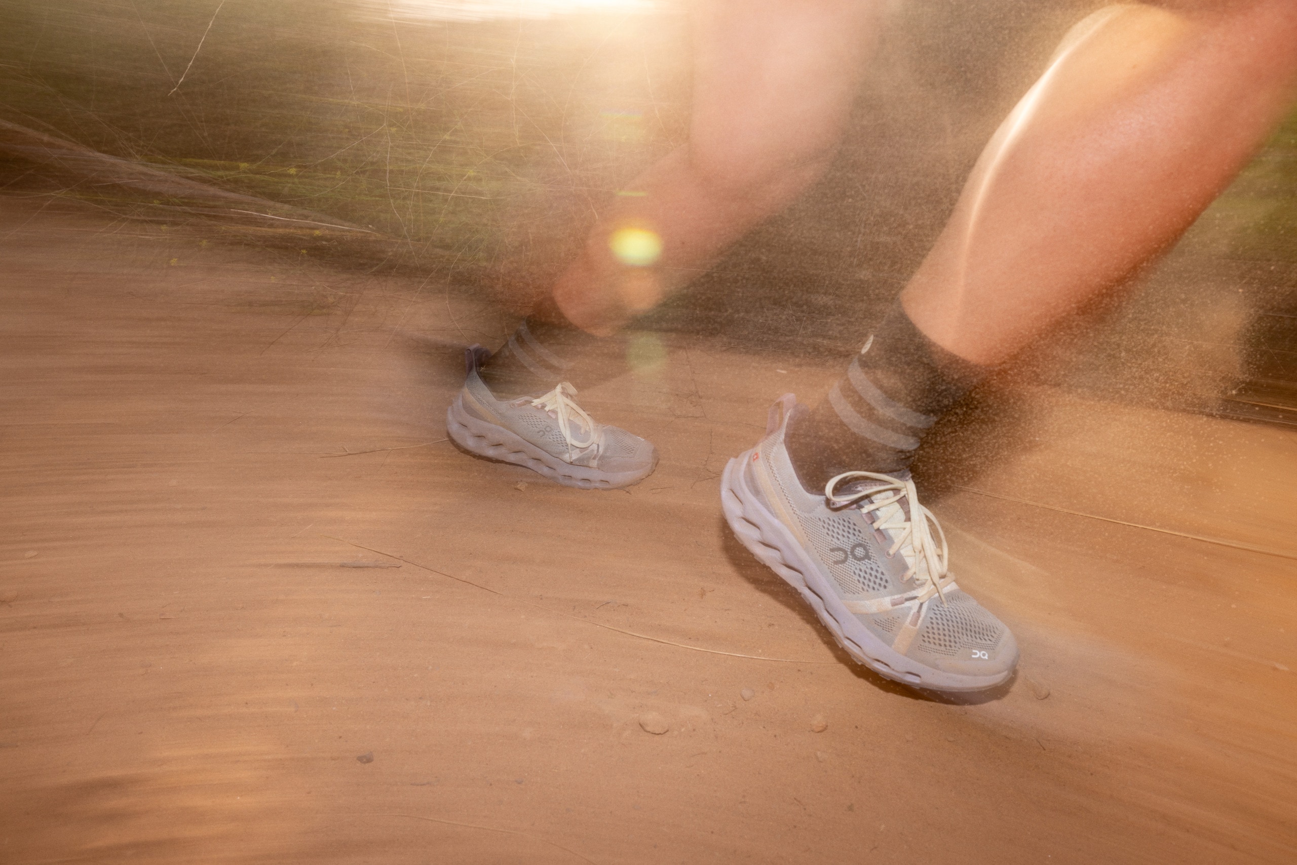 A below-the-knee view of a runner running on a trail in the On Cloudsurfer Trail trail-running shoes. The image is slightly blurred to show motion.