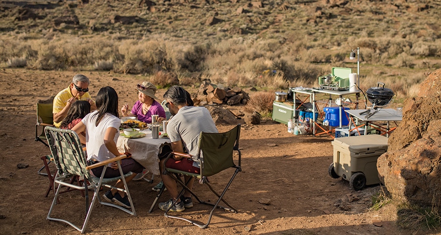 A family eats a meal at a campsite, near a homemade camp kitchen setup.
