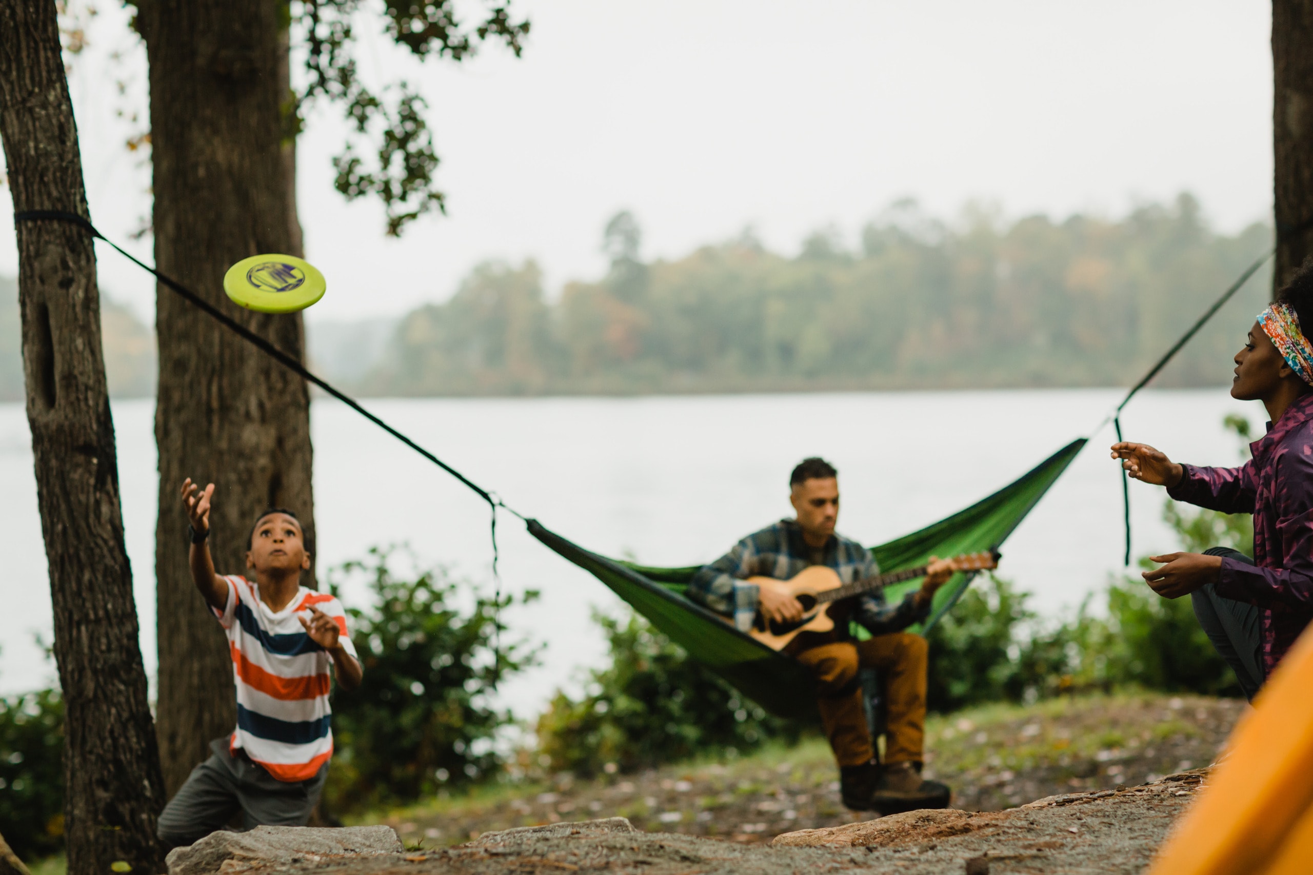 In front of a person playing guitar in a hammock is a parent and child playing with a flying disk.